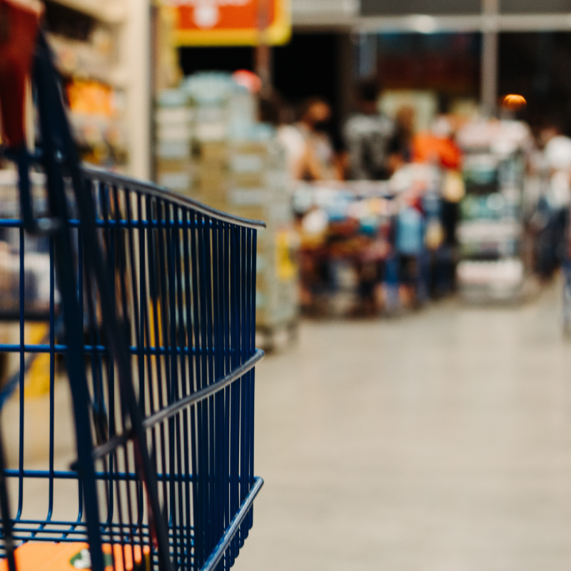 A close-up of a blue shopping cart in the aisle of a grocery store. The background is blurred, showing shelves with various products and a few shoppers. The lighting is warm, creating a welcoming atmosphere typical of bustling markets.