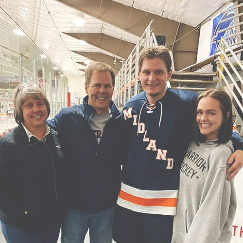 A group of four people stand together inside an ice hockey rink. One person, wearing a "Midland" hockey jersey, is flanked by Bill Finley in a casual jacket along with one older individual and a younger individual in a "Ferris State University Hockey" sweatshirt. They are all smiling.
