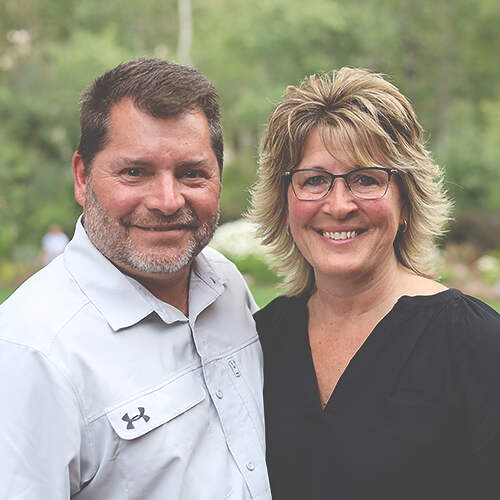 A smiling man with short dark hair, a graying beard, and a light gray button-up shirt stands next to Dana Zezzo, who has short, layered blonde hair, glasses, and a black V-neck blouse. They are outdoors with green foliage in the background.