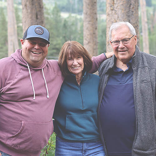 Three people, including Larry Zavadil, stand together outdoors in front of trees, smiling. The person on the left is wearing a purple hoodie and cap, the person in the middle a teal sweater, and the person on the right a grey jacket. They have their arms around each other.