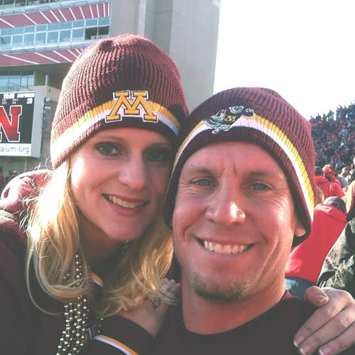 A smiling woman and man, identified as Sarah Louden and her companion, are wearing matching Minnesota beanies at a sports stadium. They are bundled up in warm clothing, and the atmosphere suggests a lively crowd in the background. The man has his arm around Sarah as they pose for the photo.