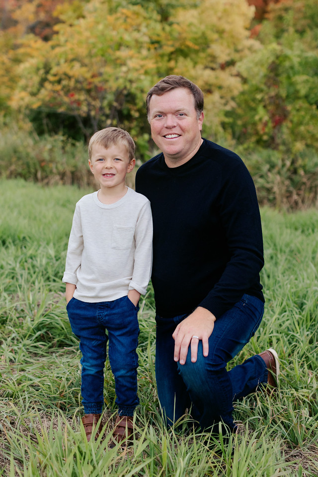 A man kneels on one knee beside a young boy standing in a grassy field. The man is wearing a black long-sleeve shirt and blue jeans, while the boy is dressed in a white long-sleeve shirt and blue jeans. Both are smiling, with a backdrop of trees in autumn colors.