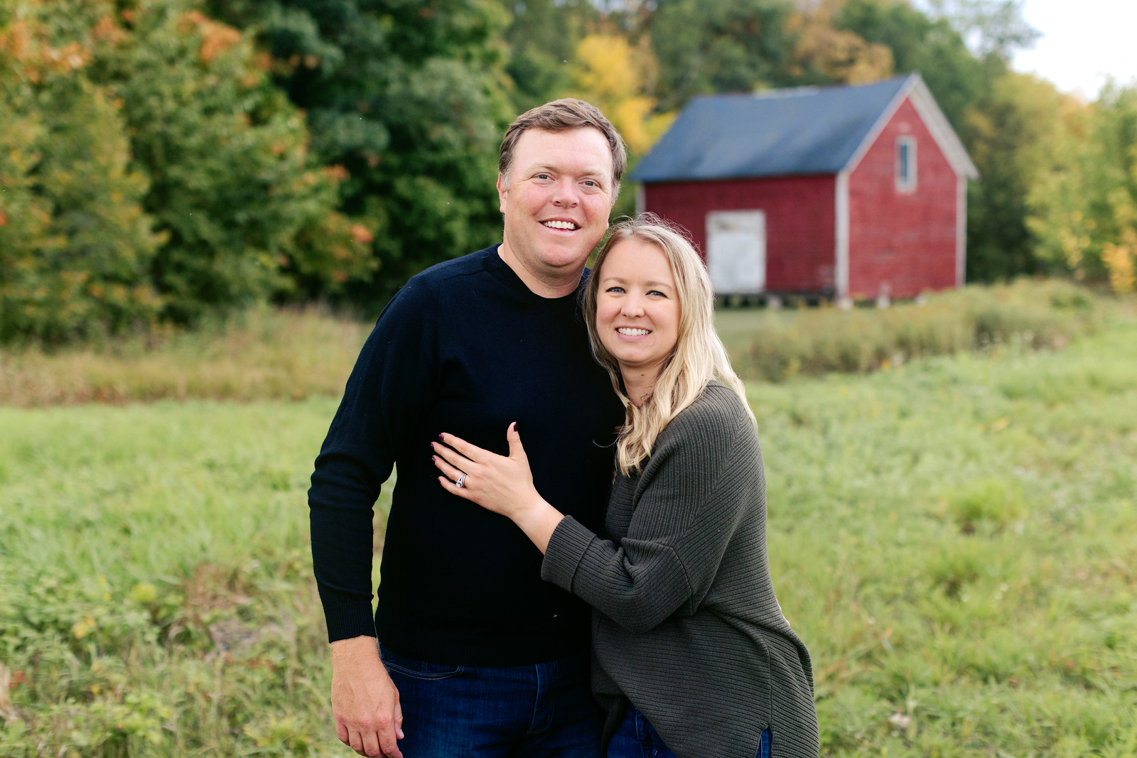 A smiling man and woman stand close together in an outdoor setting with lush greenery and a small red barn in the background. The woman has her arm around the man's waist and is showing off a ring on her hand. They both appear happy and content.