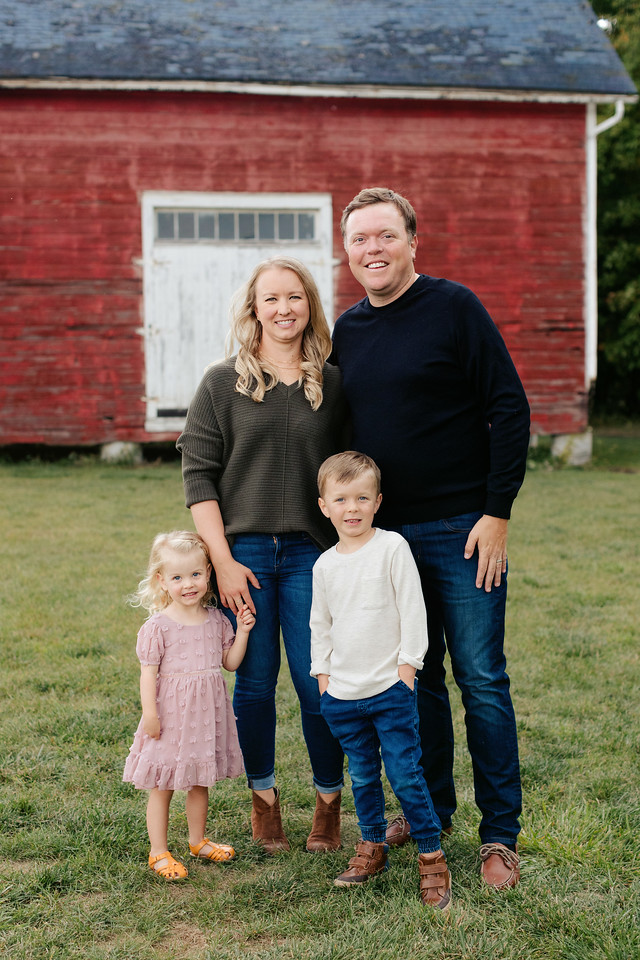 A family of four poses in front of a rustic red barn. The father and mother stand behind their young son and daughter. The father and son both wear jeans and dark tops, while the mother wears a green sweater and jeans, and the daughter wears a pink dress.