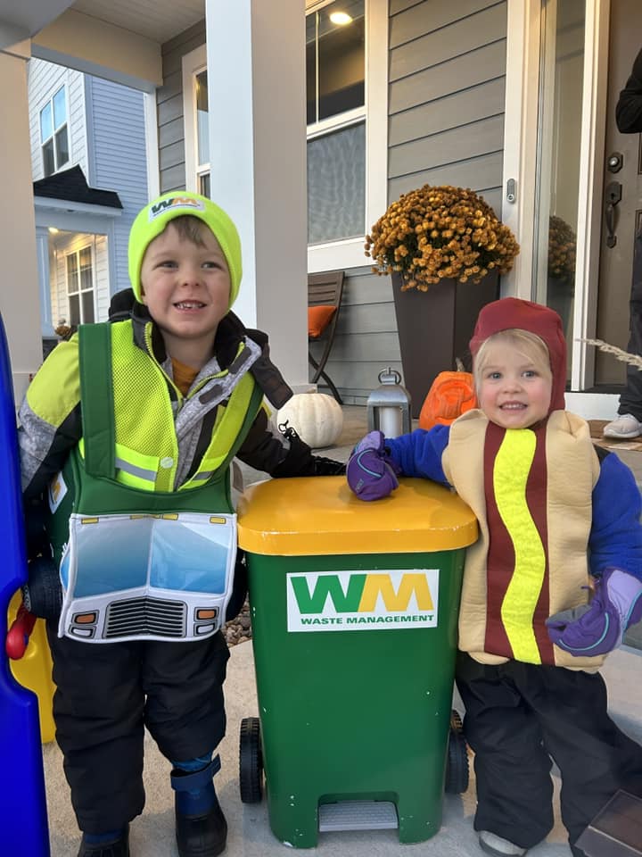 Two children in costumes stand on a porch. One is dressed as a sanitation worker with a green vest and yellow hat, next to a small green waste management bin. The other is dressed as a hotdog, smiling and holding the bin's lid. Potted flowers decorate the porch.