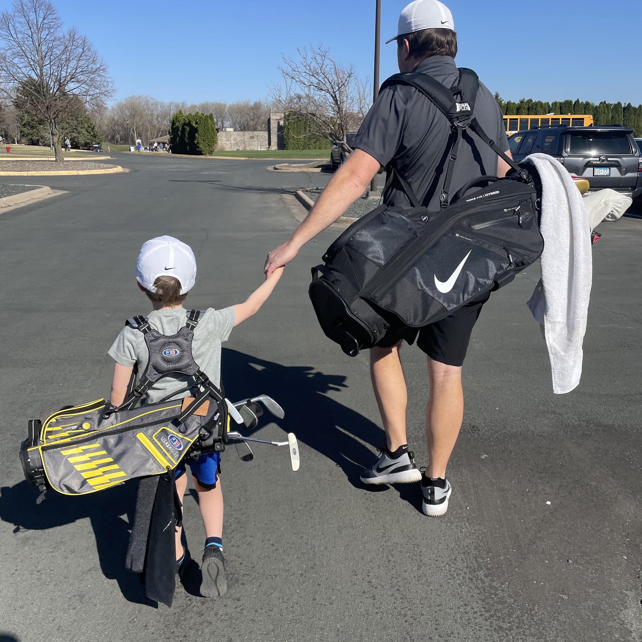 A man and a young child are walking away, holding hands. They each carry golf bags over their shoulders, with the child also carrying a putter. They both wear hats, and the setting appears to be a parking lot next to a golf course.