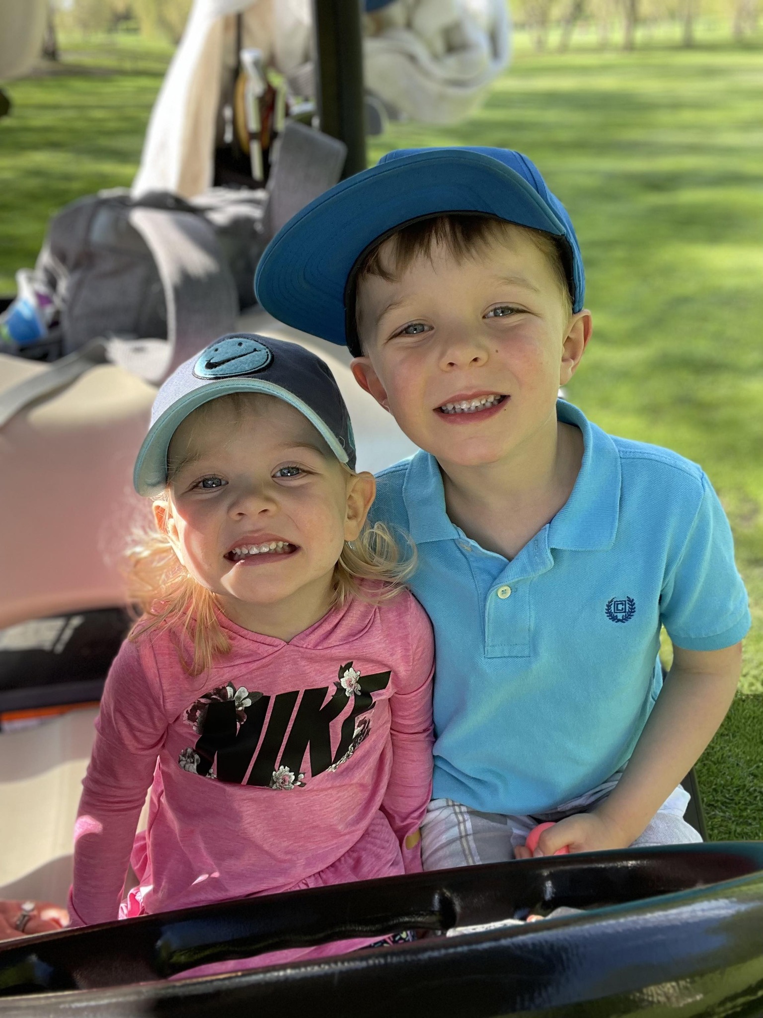 Two young children, a girl and a boy, are smiling at the camera while sitting together on a golf cart. The girl is wearing a pink Nike shirt and a blue cap, while the boy is in a light blue polo shirt and a matching blue cap. Green grass and trees are in the background.
