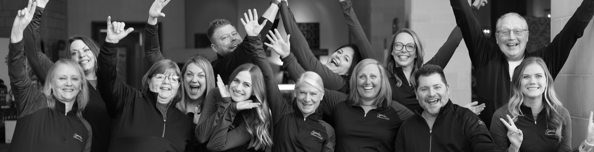 A group of individuals happily posing together, exuding a sense of culture. Some have their arms raised while others make playful gestures, all smiling and appearing joyful. They wear similar dark-colored jackets with matching logos, set against a backdrop of warm interior lighting.