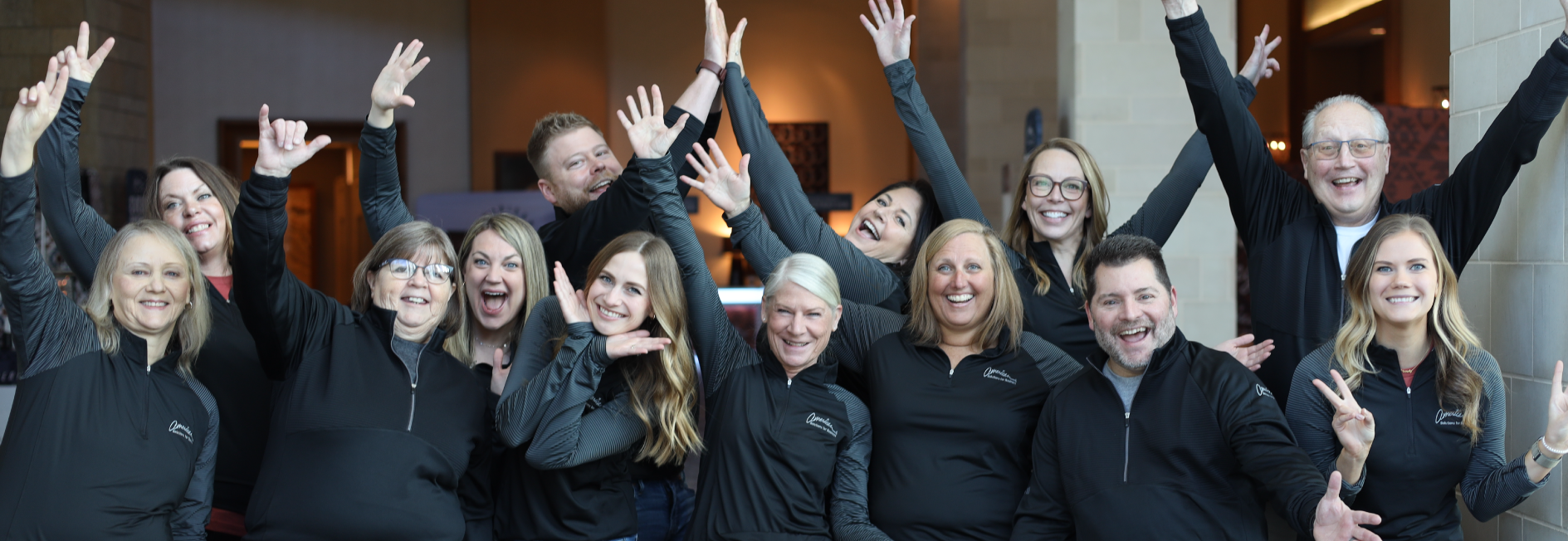 A group of 12 people, wearing matching black jackets, pose enthusiastically with arms raised and smiling faces. They are inside a building with warm lighting and modern decor, creating a joyful and energetic atmosphere that reflects their shared culture.