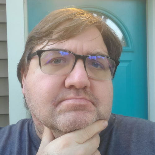 Robert Stone, a man with glasses and short brown hair, stands in front of a teal door. Wearing a gray shirt, he has his hand resting on his chin. His expression appears thoughtful.