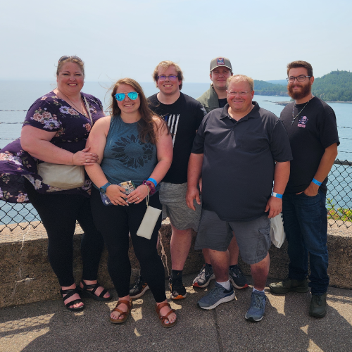 A group of six people, including Tricia Loverink, stand close together and smile at the camera. They are outdoors with a scenic view of the ocean and forested hills in the background. A chain-link fence and a concrete barrier are visible behind them.