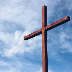 A tall, wooden cross stands against a backdrop of a blue sky with scattered, wispy clouds. The cross is the central focus of the image, symbolizing religious faith and spirituality amidst life's bustling markets. The weather appears clear and calm.