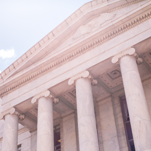 A low-angle view of a neoclassical building, featuring grand white marble columns supporting a pediment, with intricate architectural designs reminiscent of historic markets and a faint sculpture at the top, partially obscured by bright sunlight.