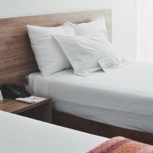 A neatly made bed with white sheets and pillows in a modern hotel room evokes a sense of calm amid the bustling city markets. A phone and notepad sit on the wooden bedside table, while a note is placed on the bed. Another bed with a red blanket is partially visible in the foreground.
