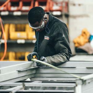 A person wearing safety glasses and ear protection is measuring a metal frame using a measuring tape in an industrial workshop, which operates in various markets. Shelves with yellow bins and various equipment are visible in the background.