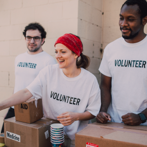 Three volunteers in white "VOLUNTEER" t-shirts stand by a table with boxes labeled "AID" and "Medicine." One person hands out colorful cups while the others stand nearby smiling. They appear to be outdoors near a building, reminiscent of bustling markets where communities come together.