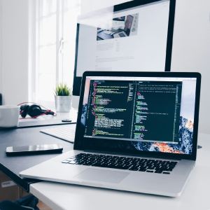 A laptop displaying colorful code on the screen sits on a white desk. Next to the laptop is a smartphone, a pair of headphones, and a cup. In the background, a large desktop monitor displays market trends on a web page. A plant and a window are also visible.