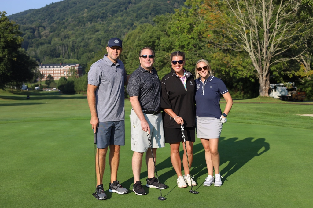 Four people pose with golf clubs on a sunny day at a green golf course. A tree-covered mountain and a large building are visible in the background, possibly the venue for the ASB Annual Summit. They are all smiling and dressed in casual golf attire.