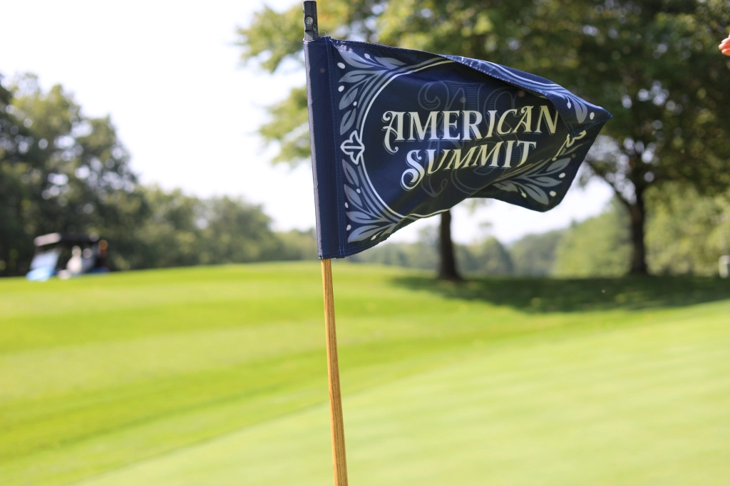 A golf course with a close-up of a flag reading "American Summit." The background showcases trees and a golf cart on a sunny day, reminiscent of the serene landscapes found in Virginia.