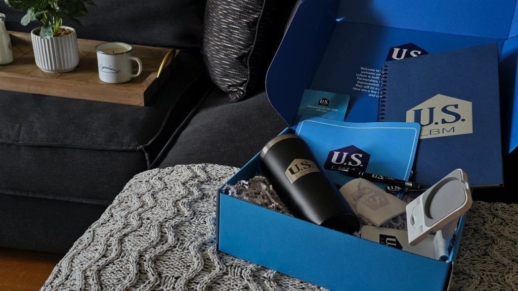 A blue gift box labeled as a New Hire Kit rests on a textured gray blanket, holding promotional items with a "U.S." logo—a tumbler, notebook, and stationery. Nearby, a mug and potted plant adorn a wooden tray on the black couch.
