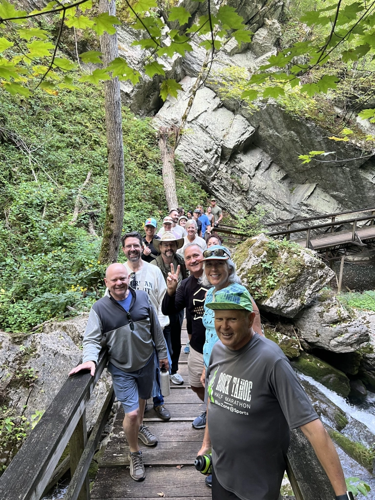 A group of people stand on a wooden bridge surrounded by lush greenery and large rocks in Virginia. Some are smiling at the camera, while a few make peace signs, enjoying their hike near the hot springs during the ASB Summit in this scenic forested area.