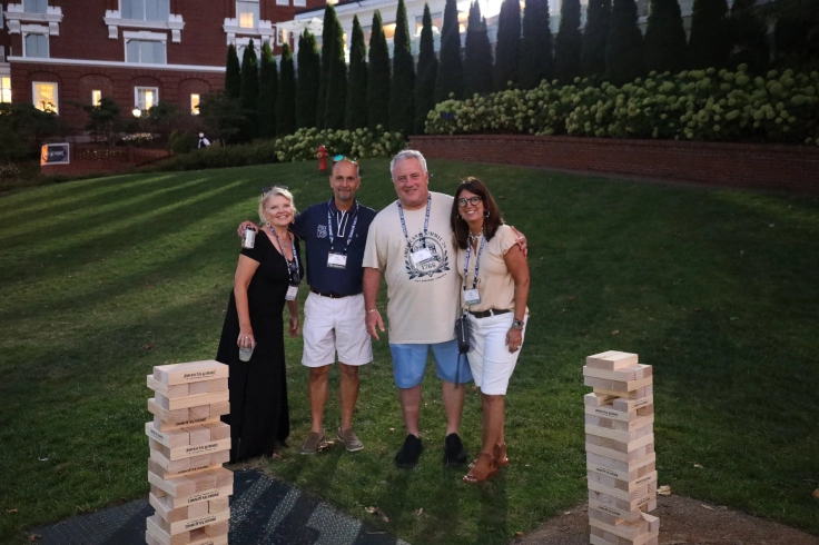 Four people stand smiling on a grassy area by two Jenga towers during the Annual Summit. They are outdoors with a backdrop of a building and greenery, embodying the spirit of ASB under the pristine sky.