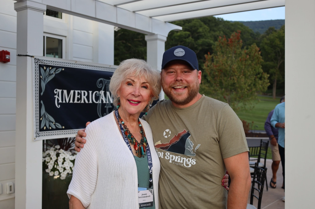 An elderly woman and a man stand closely, smiling at the camera during the annual summit. The woman wears a white cardigan and colorful necklace, while the man sports a baseball cap and t-shirt. They're outdoors with a sign reading "American" in the background.