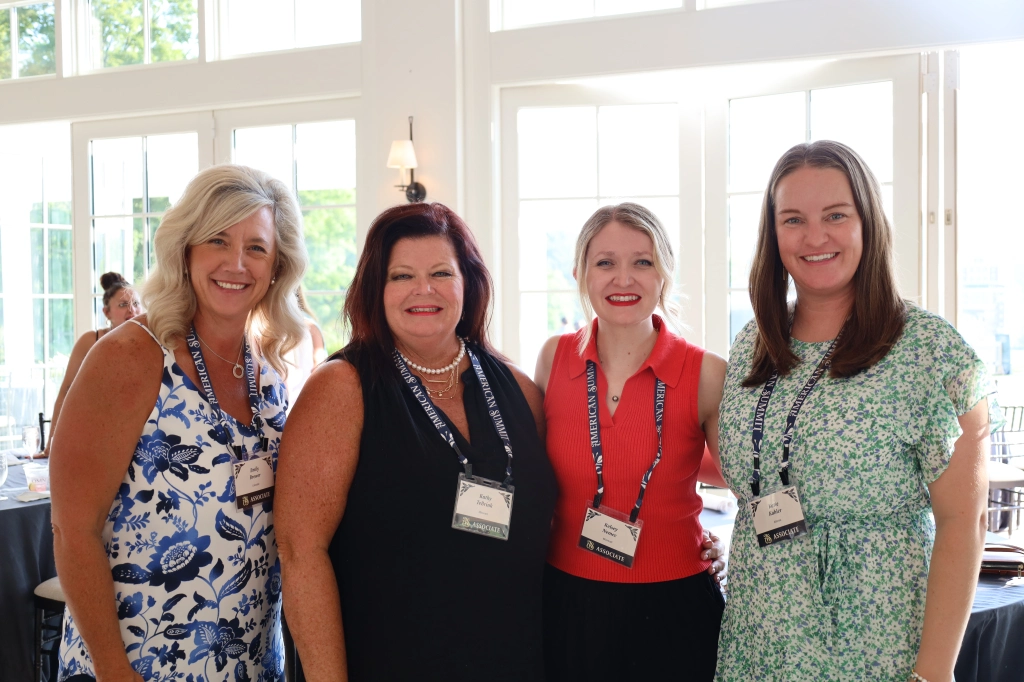 Four women stand together, smiling, in a bright room with large windows at the ASB Annual Summit in Hot Springs, Virginia. They are wearing name tags and casual business attire. The atmosphere appears friendly and professional.