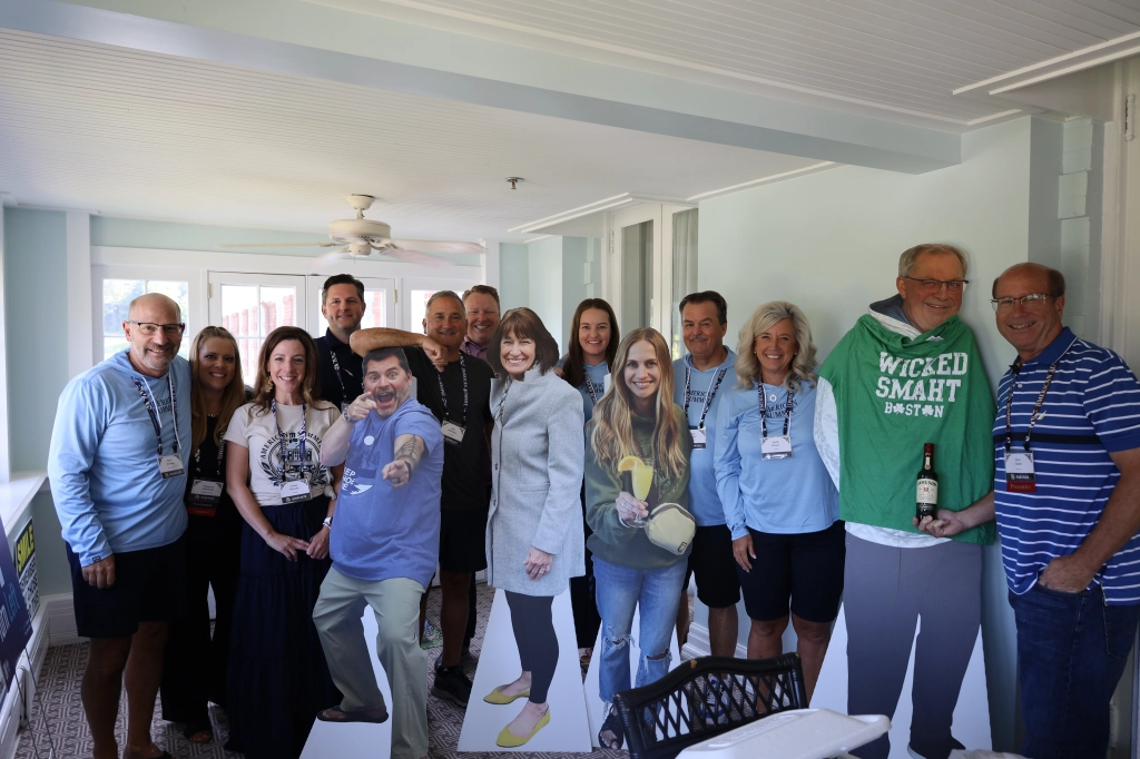 A group of people gathered indoors, smiling at the camera, some standing next to life-sized cardboard cutouts. Casually dressed, they appear to be on a bright enclosed porch, perhaps taking a break from the ASB Annual Summit.