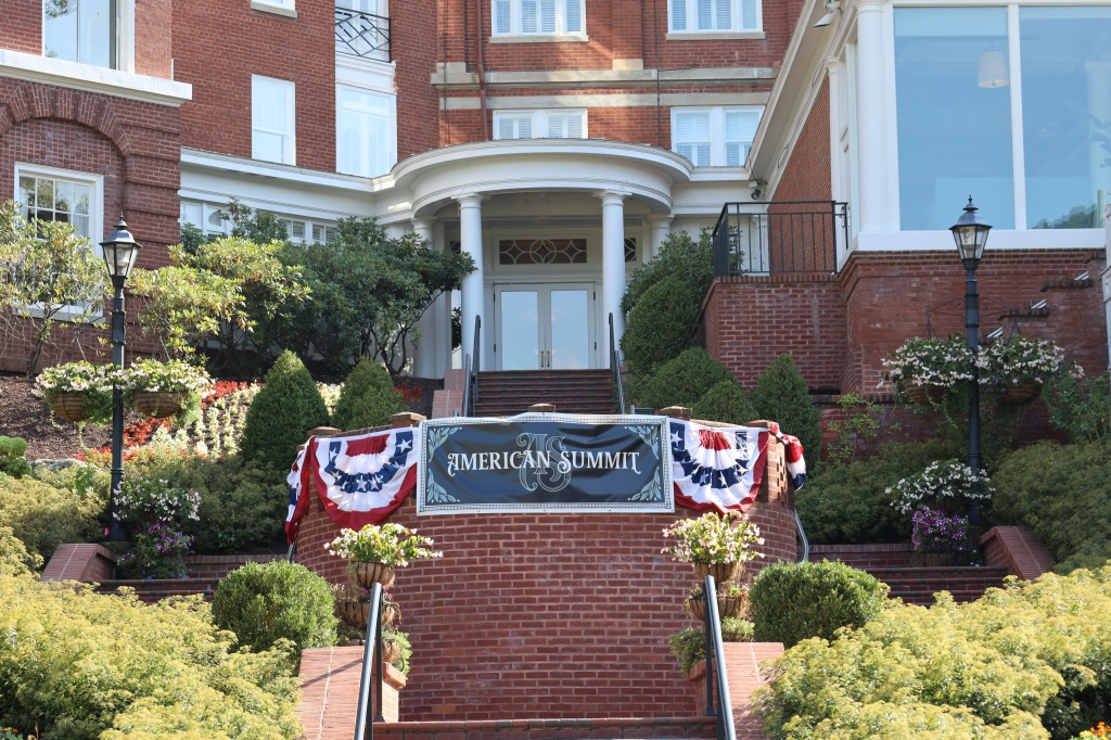 The brick building, labeled "American Summit," is decked out in red, white, and blue bunting for the Annual Summit. Located in Hot Springs, Virginia, its entrance is flanked by manicured bushes and lampposts with stairs leading up to the door.