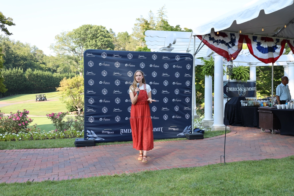 A woman in a red dress stands on a brick pathway, effortlessly speaking into a microphone at an outdoor event. Behind her is a large banner with "Auto Draft" written on it, and nearby is a tented area with tables and refreshments. The picturesque setting features lush trees and grassy expanses.