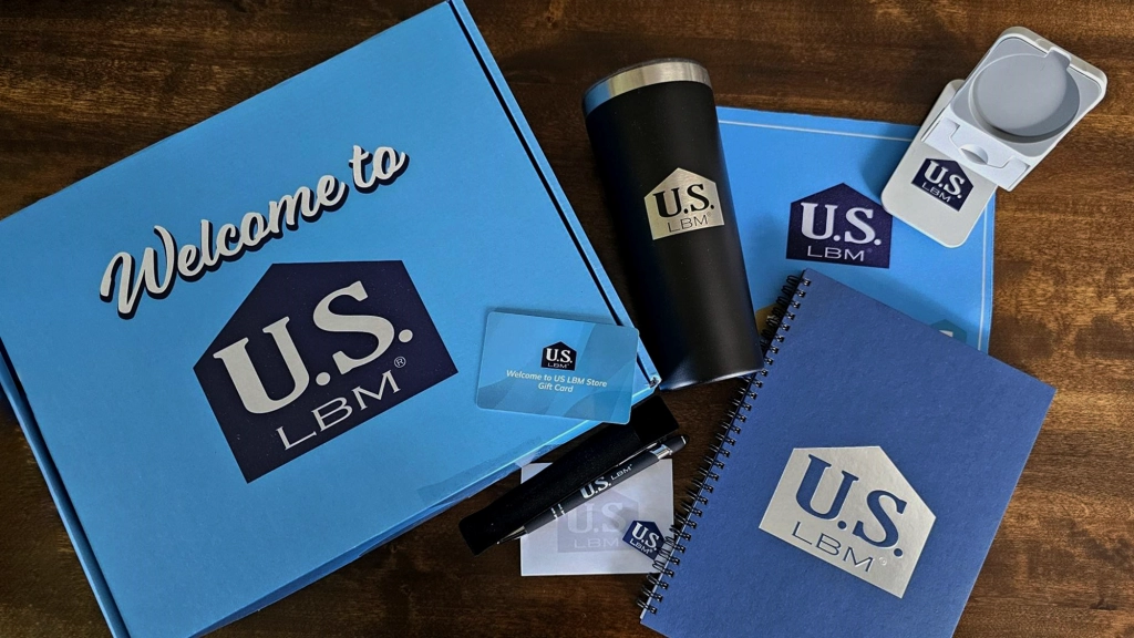 A collection of U.S. LBM branded items, perfect for new hire kits, includes a blue box with "Welcome to U.S. LBM" on it, a notebook, tumbler, business card, USB drive, and pop socket displayed on a wooden surface.