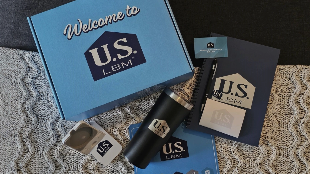 A welcome package with U.S. LBM branding, ideal for New Hire Kits, includes a blue box, a black tumbler, a notebook, earbuds in a white case, sticky notes, and a lanyard—all elegantly arranged on a textured gray and white surface.
