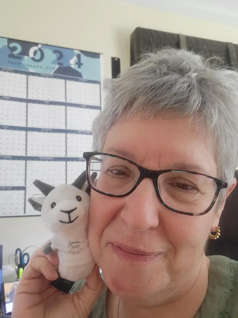 A person with short gray hair and glasses smiles while holding a small stuffed animal next to their face, perhaps a delightful piece of promotional merchandise. In the background, a 2024 calendar prominently displays August.
