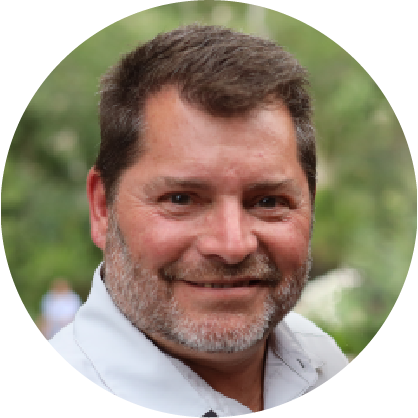 Maxx Rochette, a man with short hair and a beard, smiles at the camera. He is wearing a white shirt against a blurred outdoor setting with greenery, showcasing his connection to Canadian Solutions.