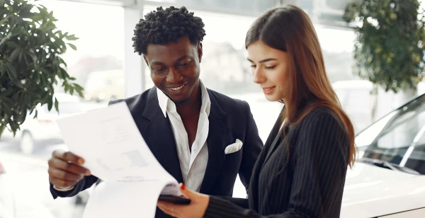 A man and a woman stand together in a bright room, examining documents. The man is smiling in his dark suit, while the woman, also in professional attire, holds the papers related to an auto draft. They are near a window with natural light filtering in, beside a plant.