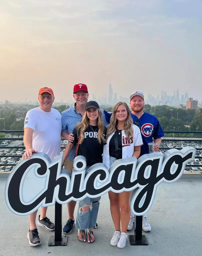 A group of five people, including Brian Deissroth, stand outdoors smiling and posing behind large letters spelling "Chicago." They're dressed casually, with some wearing baseball caps. The city skyline is visible in the background under a hazy sky.
