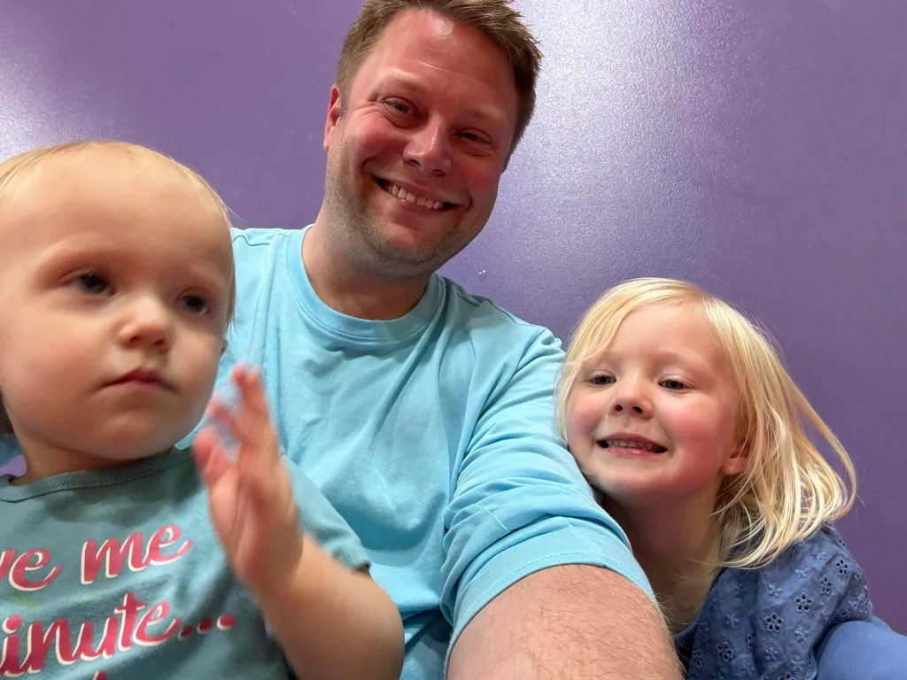 Brian Deissroth, smiling broadly, takes a selfie with two young children in front of a purple wall. The child on the left wears a gray shirt, while the child on the right, also smiling and in a blue shirt, shares his spotlight moment.