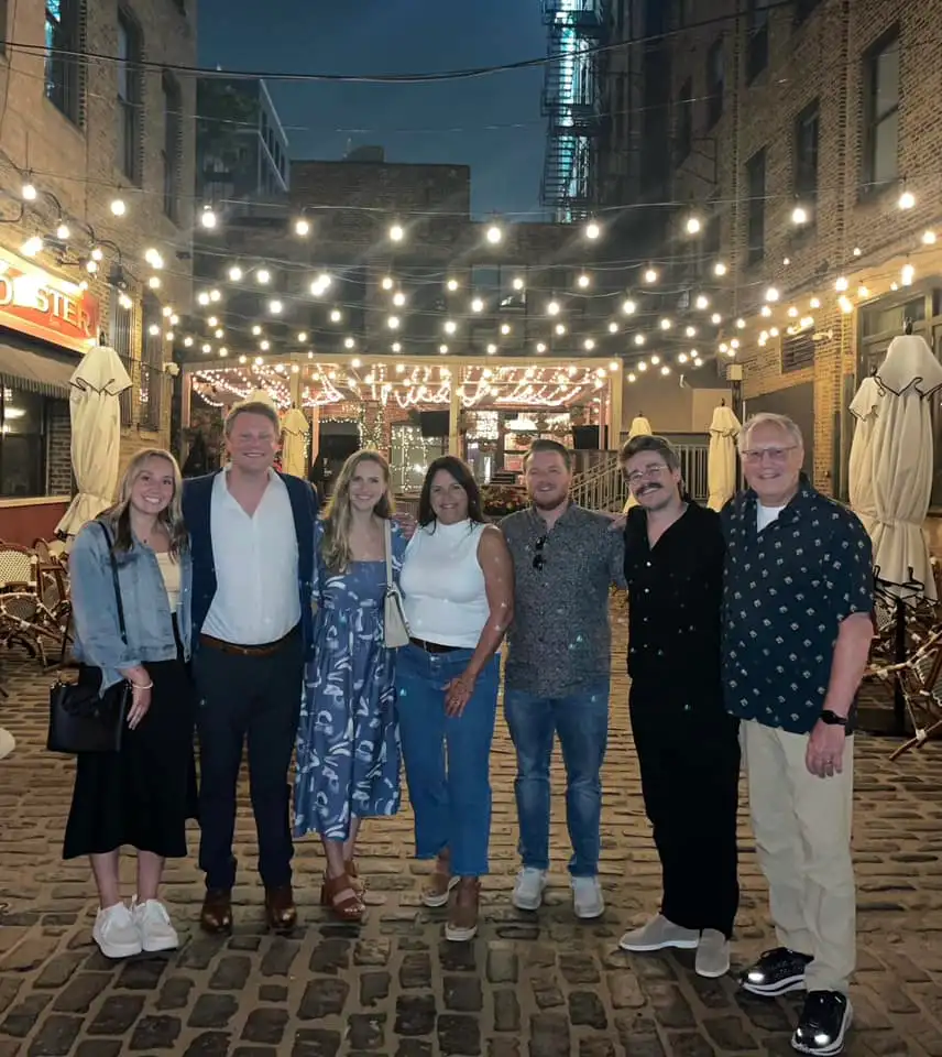 A group of seven people, including Brian Deissroth, stand closely on a cobblestone street under string lights at night. The setting is cozy, with visible outdoor seating and warm lighting creating a festive atmosphere. Some have their arms around each other, smiling in the spotlight.