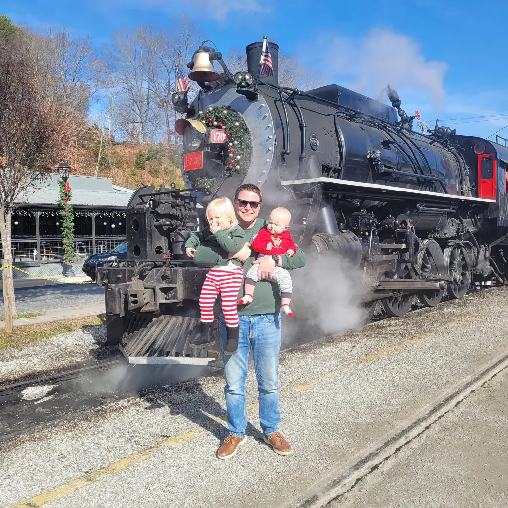 Under the holiday spotlight, a person stands in front of a vintage steam locomotive with two small children in festive outfits. The black train with red accents puffs out steam, set against trees and a building festively adorned, creating a scene reminiscent of a Brian Deissroth photograph.