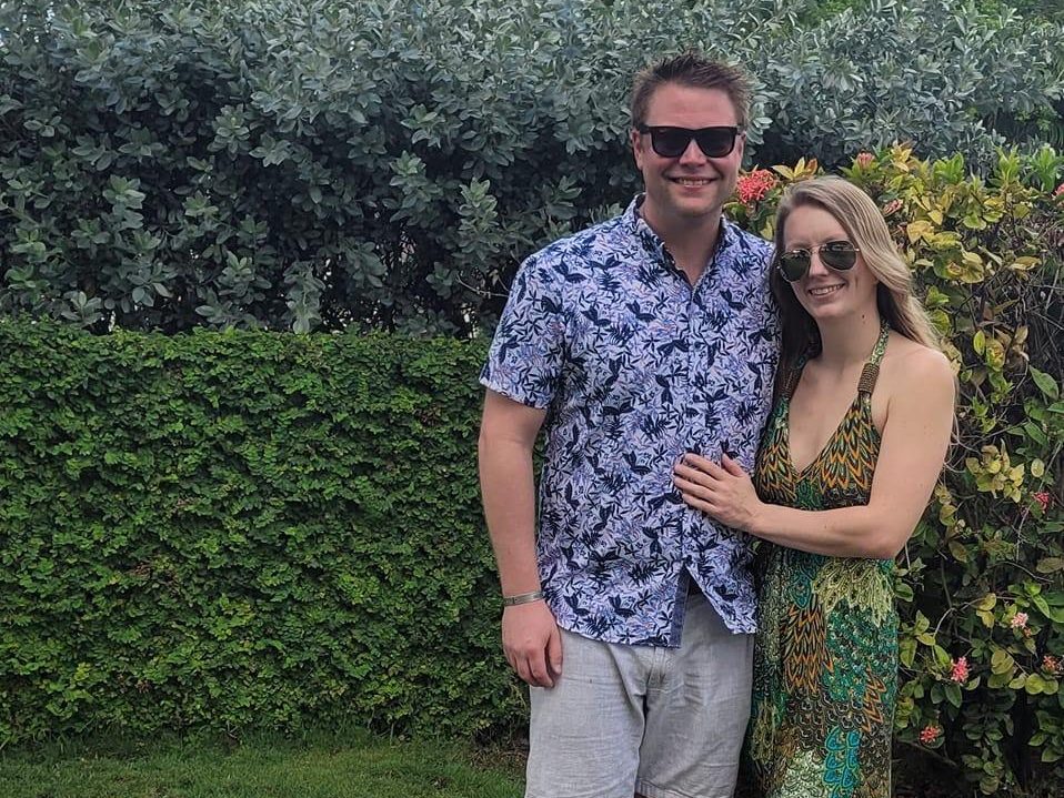 Brian Deissroth and his partner stand smiling in the spotlight of lush greenery. The man wears sunglasses, a patterned shirt, and shorts, while the woman dons sunglasses and a patterned dress. They're outdoors on a sunny day.