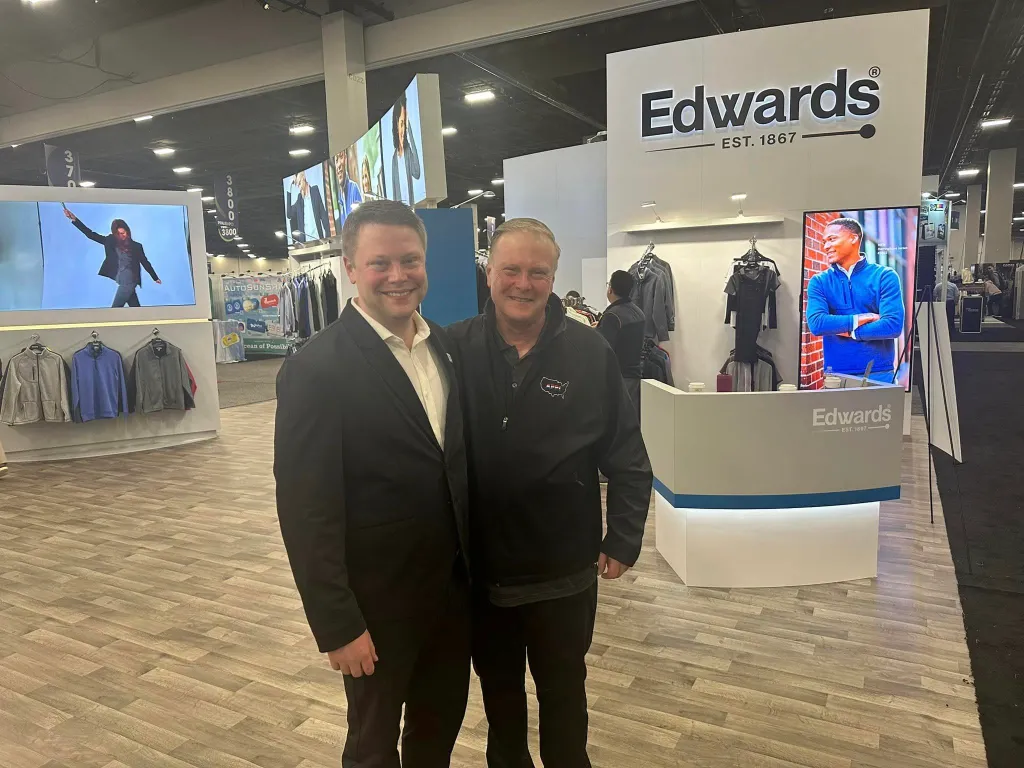 Two people, including Brian Deissroth, smile in the Edwards trade show booth. They stand on a wooden floor with clothing displays and a large screen. One wears a suit, the other a branded jacket. The booth, under the spotlight, features the Edwards logo and clothing displays prominently.