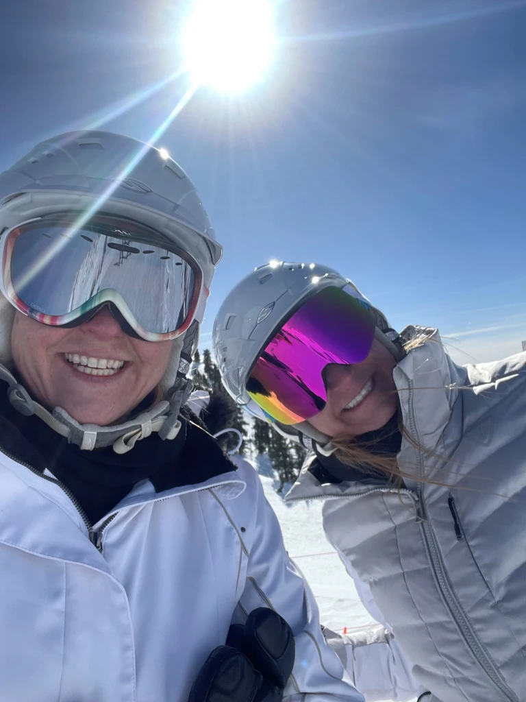 Two people in ski gear and helmets smile for a selfie on a sunny day in a snowy landscape, capturing their adventure in true Spotlight style. Their reflective goggles mirror the brilliance of the bright sun above, casting a clear blue sky as Brittany David joins them on this exhilarating journey.