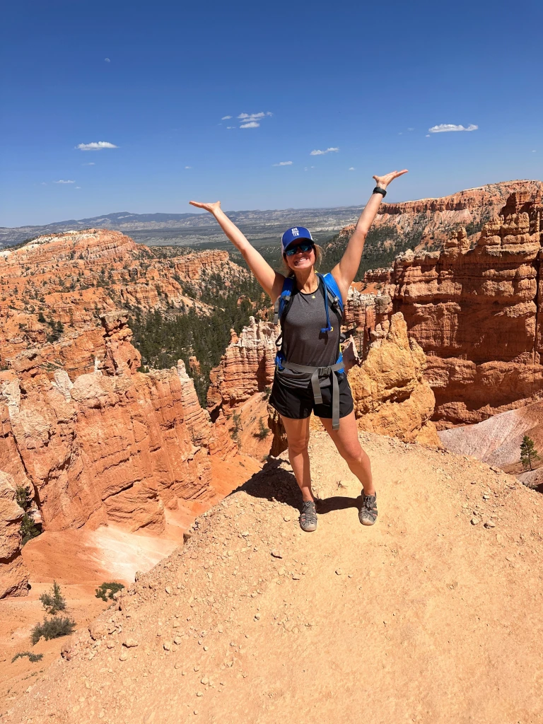 Brittany David stands on a rocky edge with arms raised, smiling. Dressed in hiking gear and a cap, she is surrounded by the orange and red rock formations of a canyon under a clear blue sky, embodying the spirit of adventure worthy of any Vendor Advisory Spotlight.