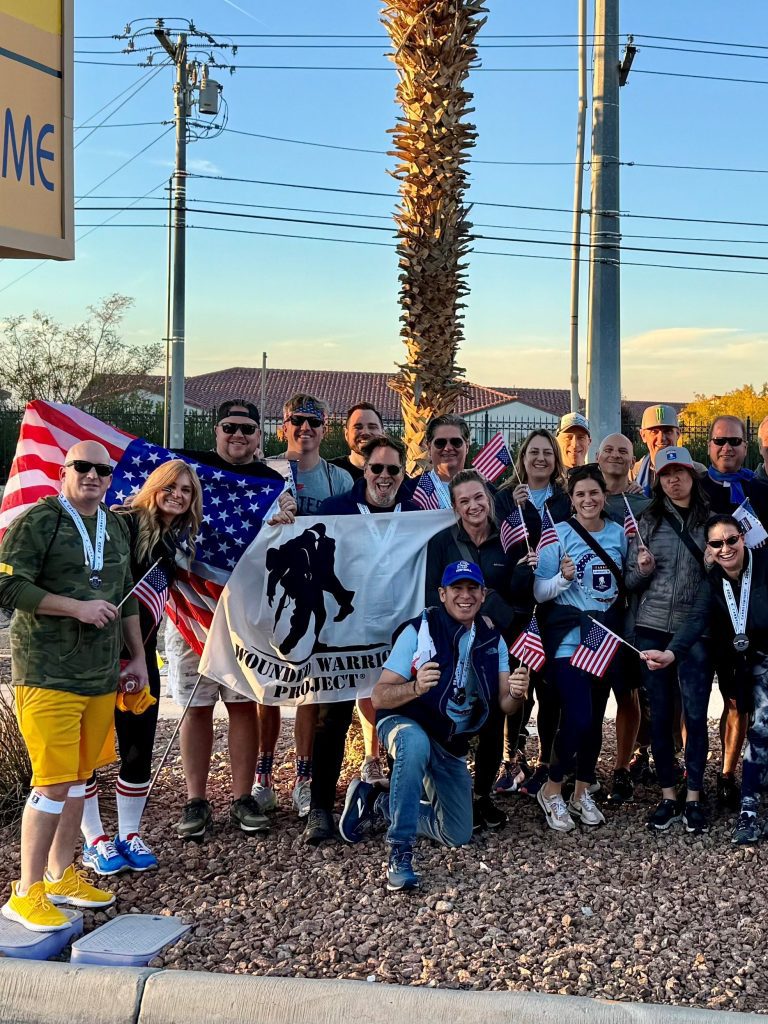 A group of people, including Brittany David, poses outdoors with a Wounded Warrior Project banner. Some hold American flags. They are casually dressed and stand on rocky ground, with palm trees and a building in the background, spotlighting their dedication to the cause.