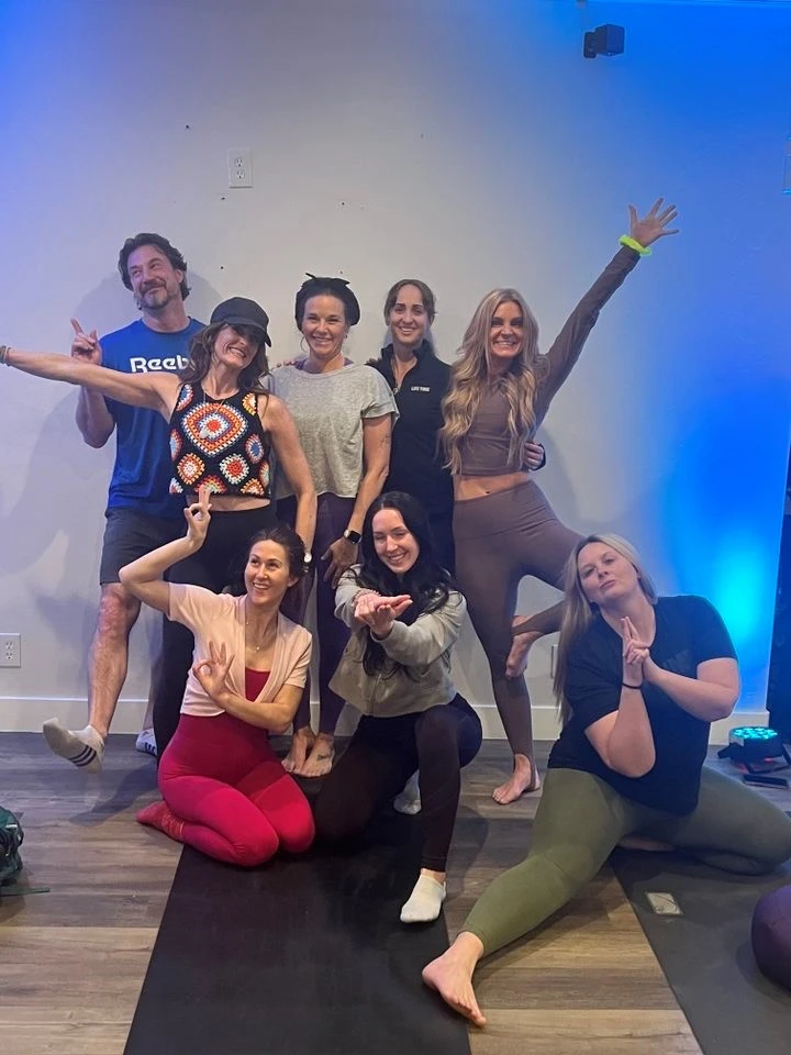 A group of eight people, including Brittany David, poses happily in a softly lit yoga studio. The blue lighting creates a relaxed atmosphere as they stand or crouch in casual and athletic attire, basking in the cheerful spotlight.