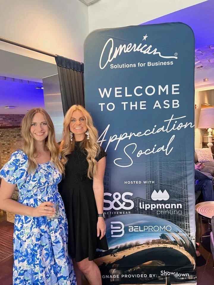 Two women smiling and standing in the spotlight next to Brittany David, in front of a banner that reads "American Solutions for Business Welcome to the ASB Appreciation Social," featuring logos for S&S Activewear, Lippmann Printing, and Belpromo.