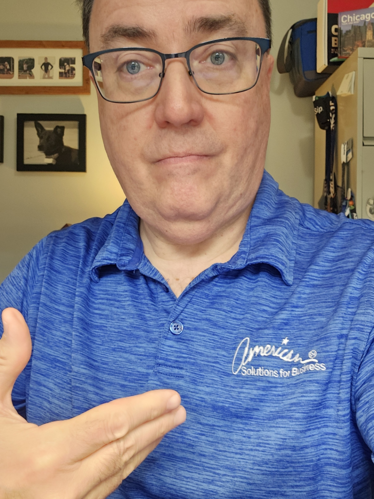 A person wearing glasses and a blue shirt with the "American Solutions for Business" logo points to it, subtly emphasizing their expertise in promotional products. The backdrop features framed pictures and a well-stocked bookshelf, setting a professional tone perfect for an August 20th campaign.