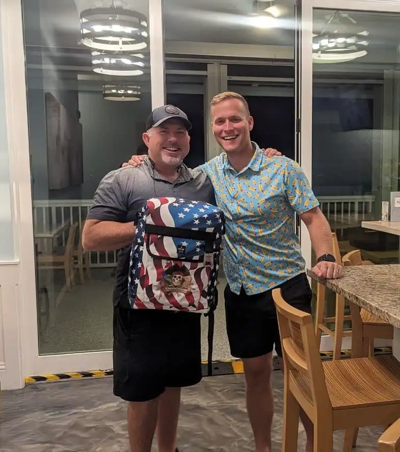 Two men smile indoors; one holds a patriotic-themed backpack, perfect for promotional products with a flag design. They stand together in a modern dining area with wooden chairs and large glass doors in the background.