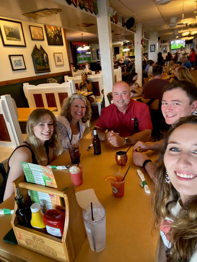 A group of five, including Vince Croyle, sit at a restaurant table, smiling and enjoying drinks. The table features condiments and a drink menu. The lively restaurant buzzes with patrons and decorations on the walls, while a TV in the background broadcasts a sports game.